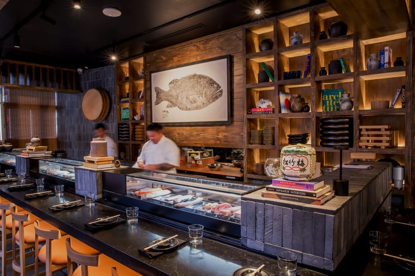 Modern Japanese sushi bar with chefs, fresh fish display, bar seating, and warm wooden decor.