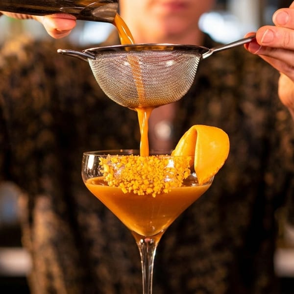 Bartender straining a vibrant orange cocktail into a coupe glass garnished with an orange peel.