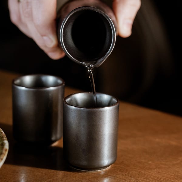 A hand pours clear liquid from a dark ceramic bottle into a small dark ceramic cup.