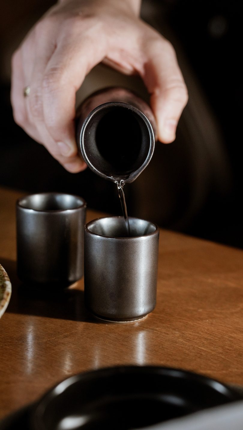Hand pouring clear liquid from a dark carafe into a small dark cup on a wooden table.