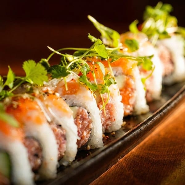 A row of sushi rolls with salmon, rice, and fresh herbs on a dark platter.