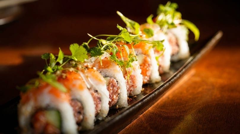 A row of sushi rolls with salmon, rice, and fresh herbs on a dark platter.