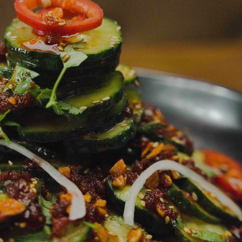 Close-up salad topped with tomato rings, cucumber slices, leafy greens, herbs, seeds, and colorful garnishes.