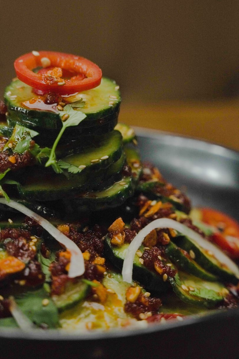 Stacked cucumber slices with chili oil, sesame seeds, red pepper, and cilantro on a dark plate.