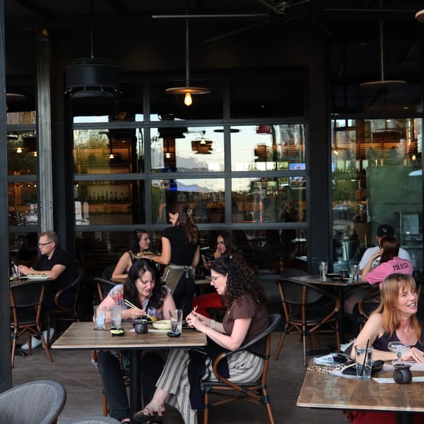 Patrons dining outdoors at a restaurant, with a server attending to tables and large windows reflecting the sky.