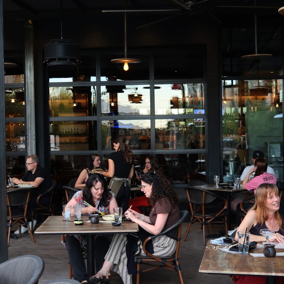 Busy restaurant patio with diners seated at wooden tables beneath pendant lights and ceiling fans.