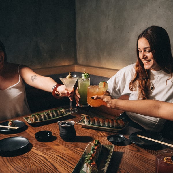 Three women toasting cocktails at a restaurant table with sushi plates.