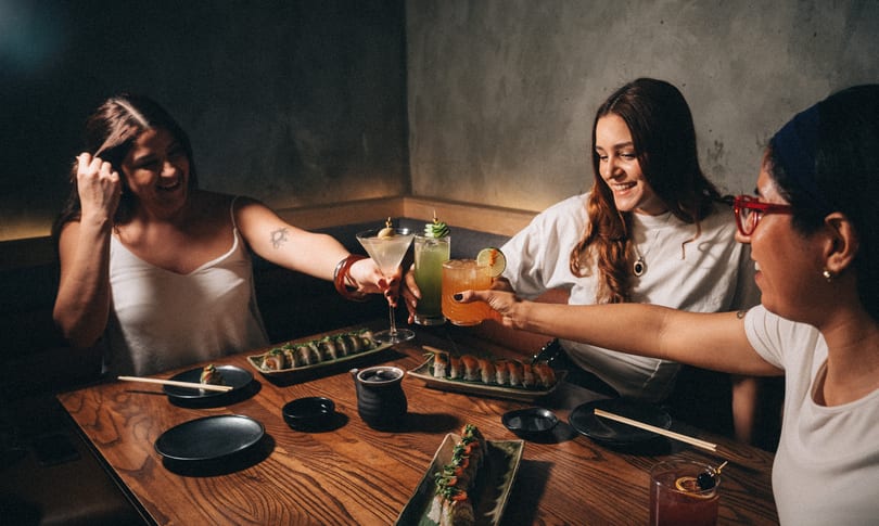Three smiling women toast cocktails at a restaurant table filled with sushi.