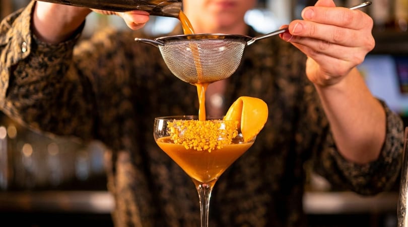 Bartender straining a vibrant orange cocktail into a coupe glass garnished with an orange peel.