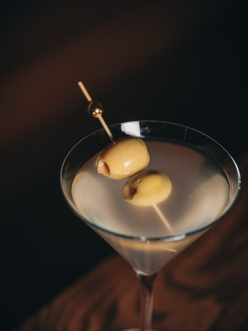 Martini glass with two green olives on a skewer against a dark background.