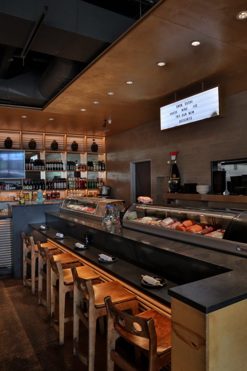 Empty sushi bar with wooden stools, a display case of fresh fish, and a lighted menu board.