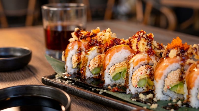 Sushi rolls with salmon, avocado, and sauce on a dark plate with a drink.