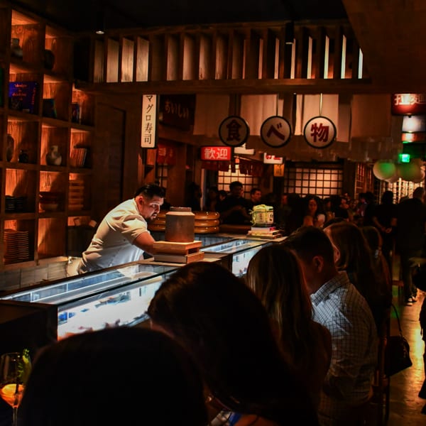 A sushi chef prepares food behind a counter for customers seated at a dimly lit bar.
