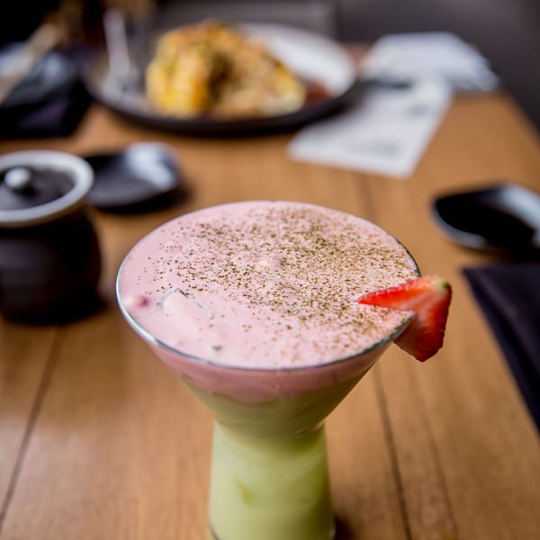 A layered green and pink cocktail with a strawberry garnish and sprinkled topping on a wooden table.