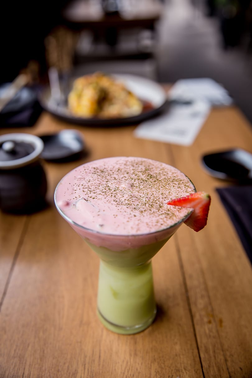 Martini glass with a green and pink layered drink, topped with powder and a strawberry slice.