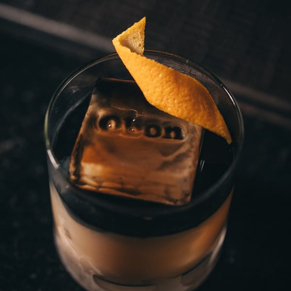 A dark cocktail with a large ice cube embossed with "Okon" and an orange peel garnish.
