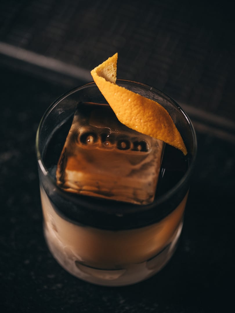 Cocktail with branded ice cube and orange peel garnish in a rocks glass.