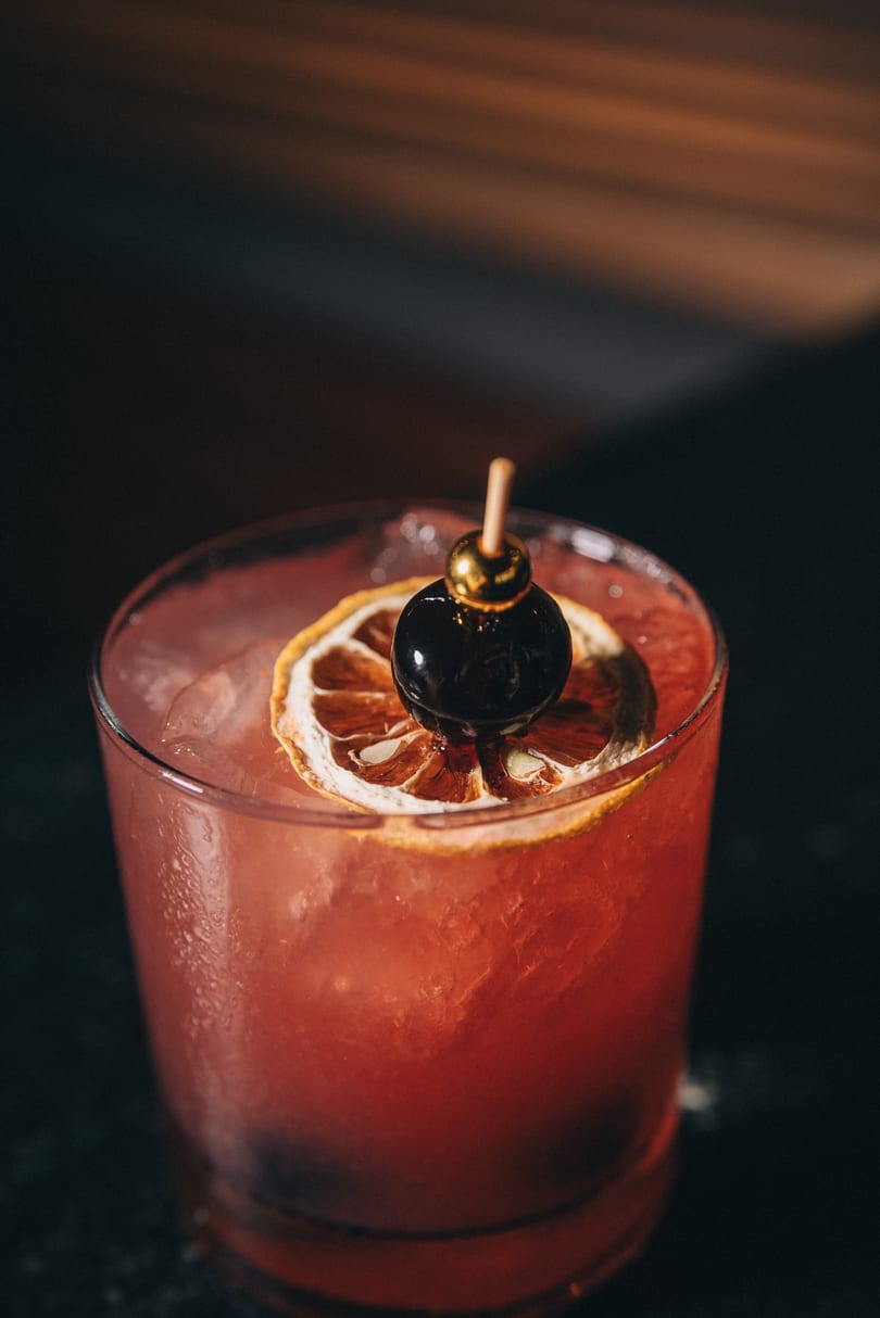 Close-up of a vibrant red cocktail with a dried orange and a cherry garnish.