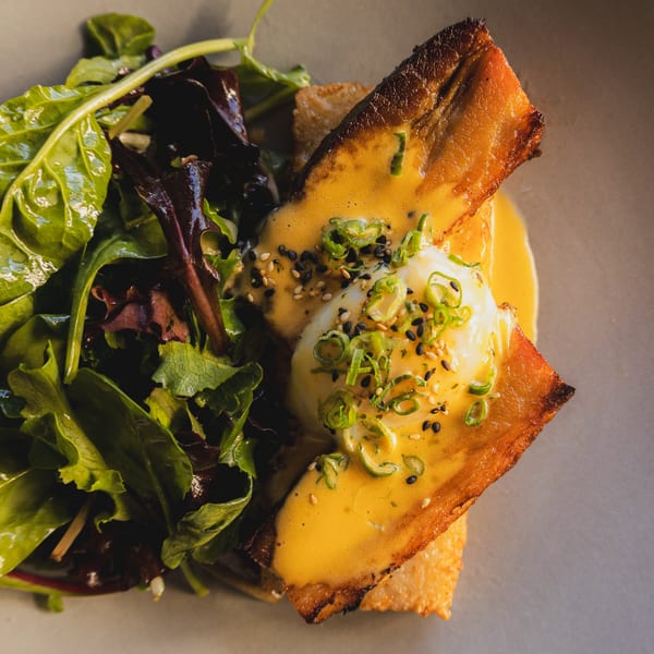 Eggs Benedict with crispy pork belly, hollandaise sauce, and a side salad on a gray plate.
