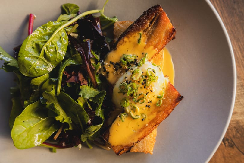 Brunch dish featuring crispy seared protein, poached egg, hollandaise sauce, and fresh greens.