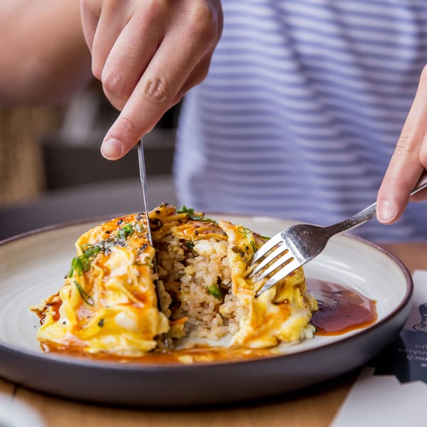 Hands cutting into an omurice dish, revealing fried rice inside, on a white and brown plate.