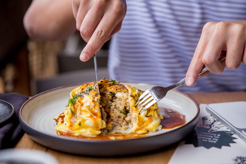 Hands cutting into omurice, revealing fried rice, garnished with sauce and sesame seeds.
