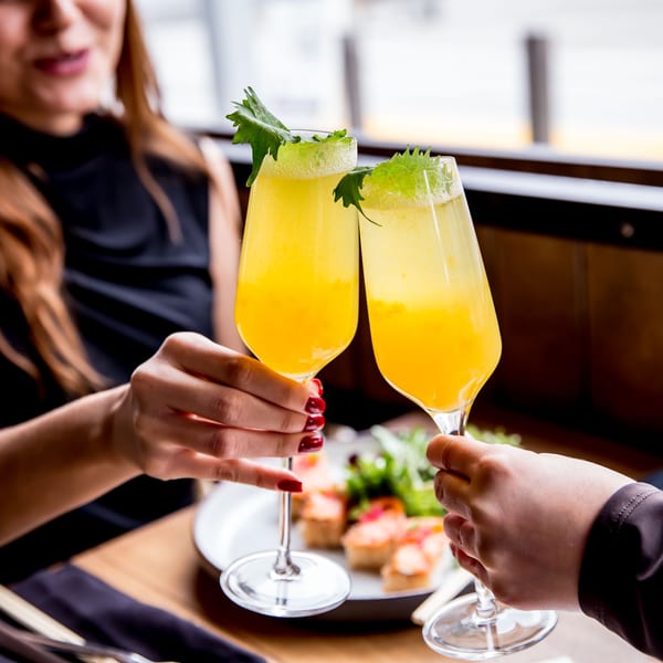 Two people clinking glasses of yellow cocktails garnished with green leaves over a plate of food.