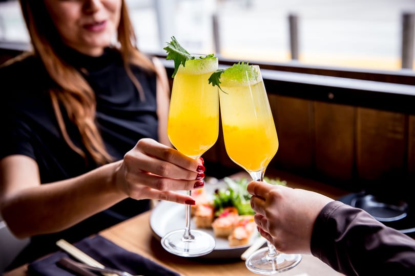 Two hands clink glasses of yellow cocktails with green garnish over a restaurant table with food.