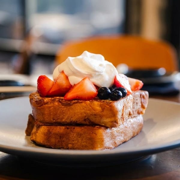 Two slices of French toast topped with whipped cream, sliced strawberries, and blueberries on a white plate.