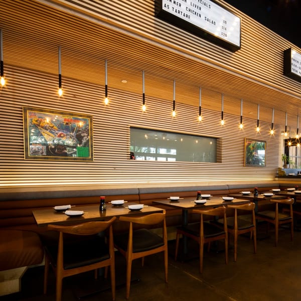 Modern restaurant interior with wooden slat walls, booth seating, individual tables, and hanging pendant lights.