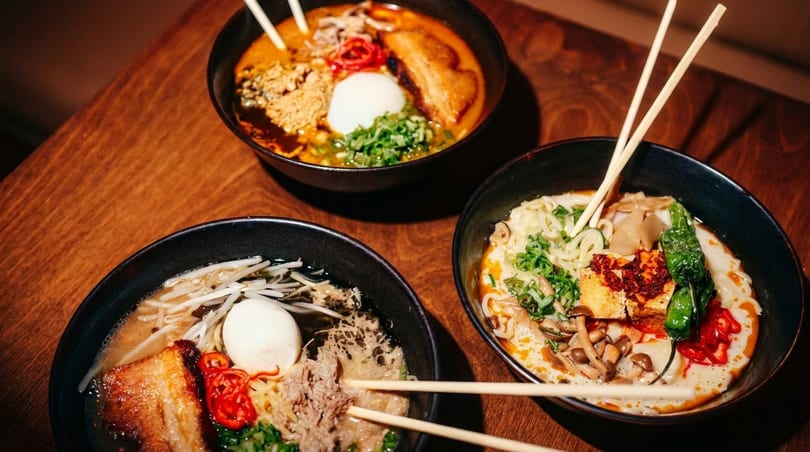 Three bowls of ramen with various toppings, including pork belly, egg, and vegetables, on a wooden table.